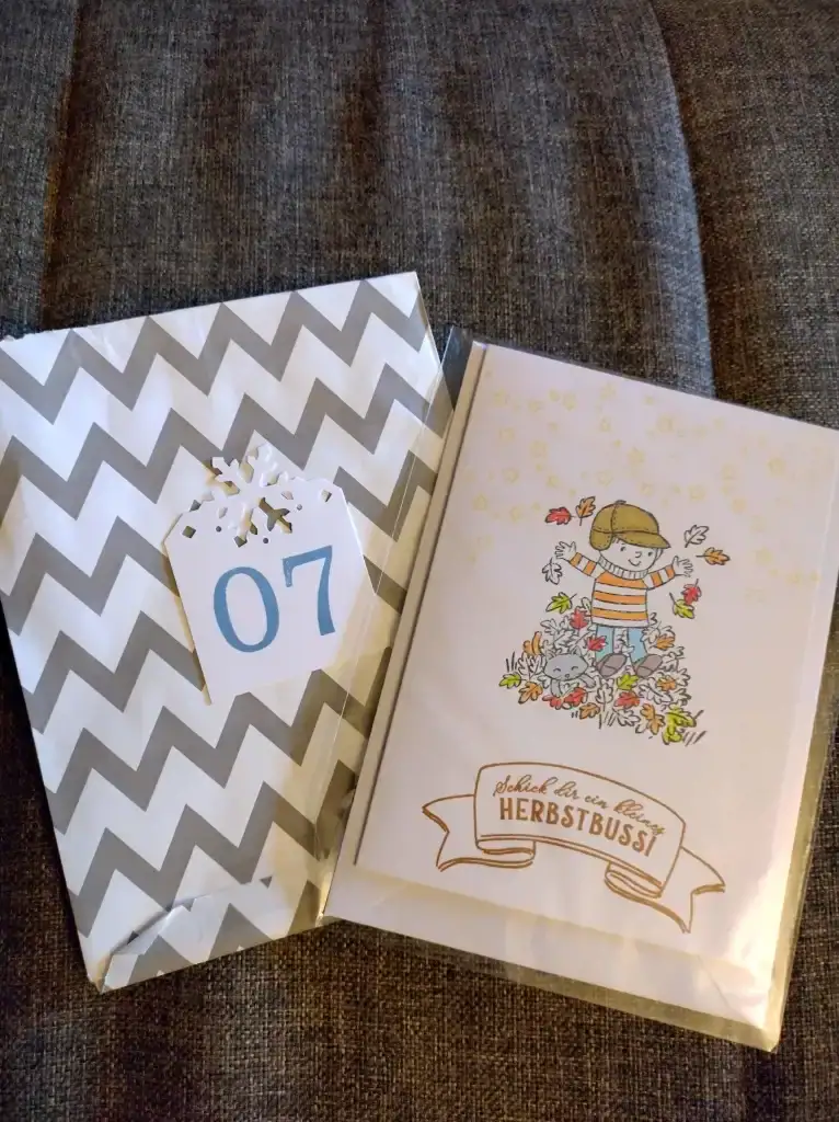 Kreativprojekte Adventskalender Karte mit einem spielenden Kind in einem Blätterhaufen. Dazu der Spruch "Schick dir ein kleines Herbstbussi". Gestaltet von Petra